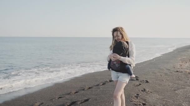 Мать держит своего ребенка на повязке и ходит вдоль морского побережья — стоковое видео