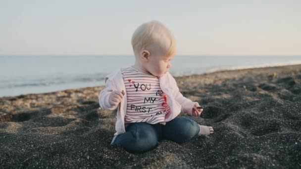 Ежегодная девушка в розовой куртке показывает камень с пляжа у моря — стоковое видео