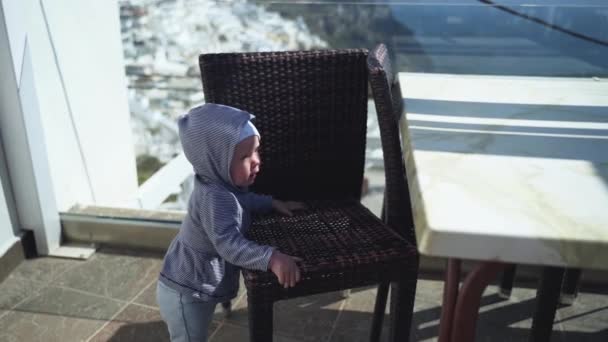 Маленький ребенок стоит и держит стул на террасе рядом со стеклянным забором, вид на остров Санторини — стоковое видео