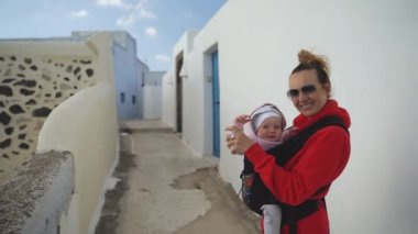 Bir Sling içinde mutlu bir anne ve kızı güneşli bir gün, kırmızı bir kazak bir kadın sokakta yürürken, bir çocuk bir hoodie giyinmiş, Santorini sokaklarında
