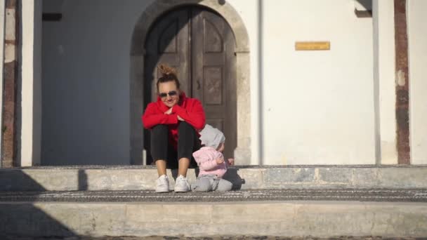 Um bebê rasteja na rua, a mãe se senta ao lado dela e cuida de sua filha, uma garotinha fofa rasteja e sorri no chão em uma rua aconchegante ensolarada, um bebê marco — Vídeo de Stock