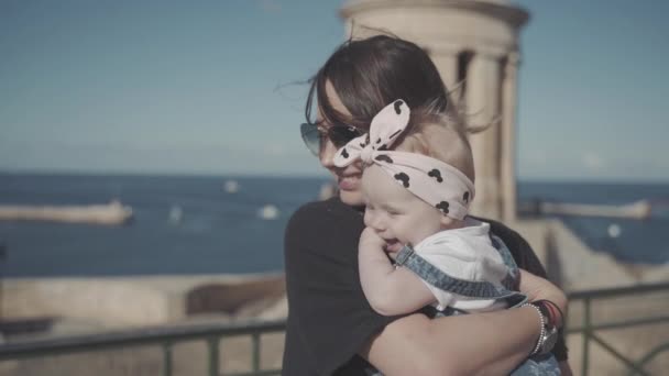 Un plan médian avec un effet de gris d'une femme debout dans les rayons du soleil avec son bébé près de la côte de la mer dans une ville La Valette — Video