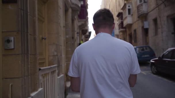 Un plan intermédiaire de l'arrière d'un homme marchant dans les rues étroites typiques avec des balcons colorés à La Valette, Malte — Video