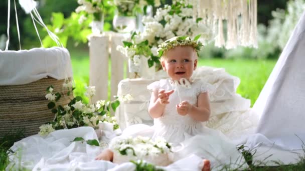 Ένα μικρό μωρό με το λευκό φόρεμα και ανθισμένο στεφάνι κάθεται στη διακόσμηση και τα χαμόγελα. — Αρχείο Βίντεο