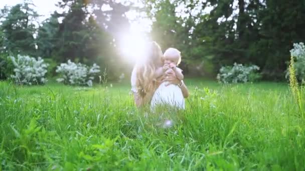 Een jonge moeder kusjes de baby in zonnestralen in het groene park. Slow Motion. Lens flare, buiten schieten. Portret. Feestsfeer. — Stockvideo