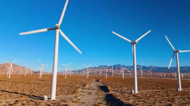 Dinamik insansız hava aracı, Palm Springs, Kaliforniya 'nın çöl manzarasındaki yüksek rüzgar türbinlerinin altından alçak bir açıdan yükseliyor. Kamera düzgünce yükselir, ölçeği ve gücü vurgular.