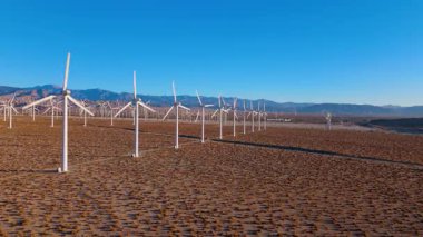 Palm Springs, California yakınlarındaki bir çöl rüzgar çiftliğinin üzerinden uçmak. Kamera, uzun rüzgar türbinlerinden oluşan simetrik bir çizgiye paralel süzülür. Kurak arazi boyunca uzanır ve dağlar uzaktadır.