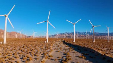 Palm Springs, California yakınlarındaki kurak çöldeki bir rüzgar çiftliğinden ileri doğru yumuşak bir hava aracı hareketi. Görüntüler, kumlu arazide yavaşça dönen uzun beyaz rüzgar türbinlerini yakalıyor.