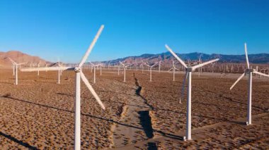 Yakın mesafe drone görüntüleri Palm Springs, Kaliforniya yakınlarındaki bir çöl rüzgâr tarlasında geniş rüzgar türbinlerini sorunsuzca geçiyor. Kamera dönen pervaneler arasında süzülüyor.