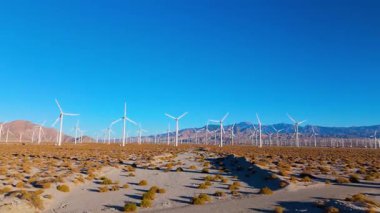 İnsansız hava aracı bir çöl vadisinde kurulmuş geniş bir rüzgar türbini tarlasında hızla ilerler. Parlak güneş ışığı ve minimum manzara temiz enerjiyi ve endüstriyel ölçeği vurguluyor. Çevre için ideal
