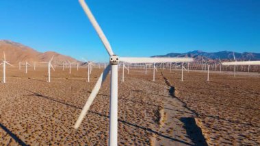 Bir rüzgâr türbini, dönen bıçaklarını keskin detaylarıyla bir çöl manzarasına ve uzak dağlara karşı yakalıyor. Dinamik perspektif temiz enerji, hareket ve modern mühendisliği vurgular. İdeal
