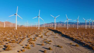 İnsansız hava aracı, kurak bir çölde geniş bir rüzgar çiftliğinin üzerinde yavaşça dönen türbinlere doğru süzülüyor. Sahne ölçeği, çevresel yenilik ve temiz enerji üretimini parlak bir ışık altında iletir.