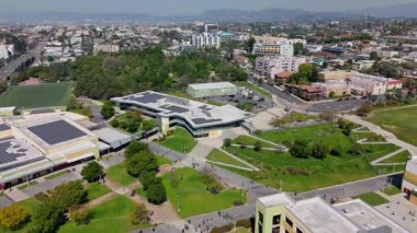 Drone footage capturing a forward flight over a modern educational complex surrounded by green park areas and urban residential buildings in Los Angeles. The camera moves smoothly forward, revealing