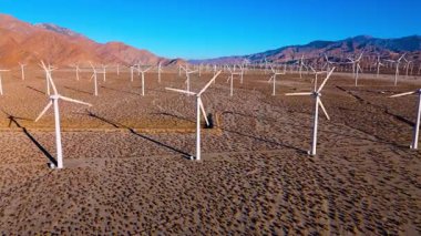 Aerial shot flying over rows of rotating wind turbines in the Palm Springs desert during sunrise. Clean energy infrastructure stretches toward the mountains under clear skies, symbolizing progress and