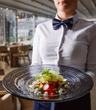 Garson elinde turp, beyaz peynir ve domatesli sebze salatası tutuyor.