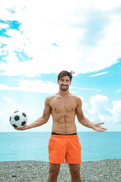 Sportif, koyu renk saçlı, turuncu şortlu, kumsalda futbol oynayan adam.