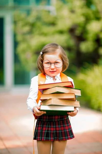 a small beautiful girl with glasses and a school uniform with books is standing near the school