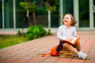 Okul üniformalı ve gözlüklü küçük bir kız, okulun yakınındaki bir yığın kitabın üzerinde oturuyor.