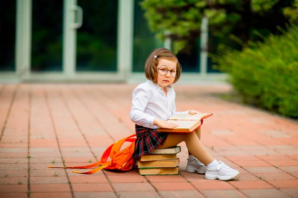 一个穿着校服和眼镜的小女孩 坐在学校附近的一堆书上 — 图库照片