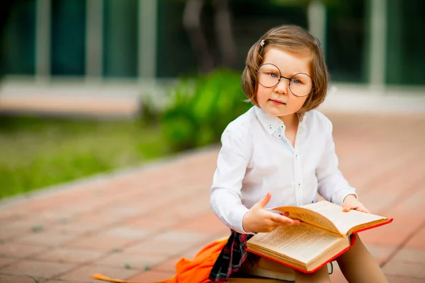 a little girl in a school uniform and glasses,sitting on a stack of books near the school