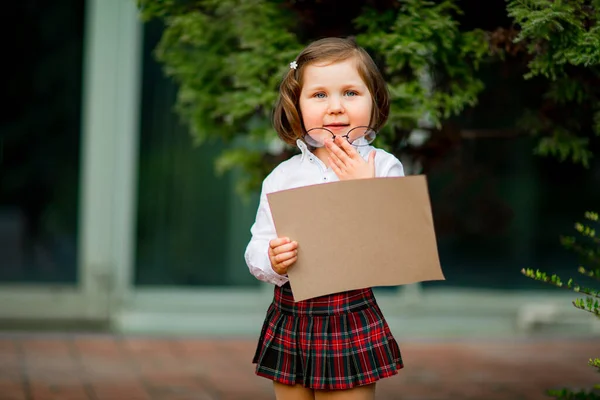 En flicka i skoluniform står nära skolan, med en bit papper, en layout med plats för text — Stockfoto