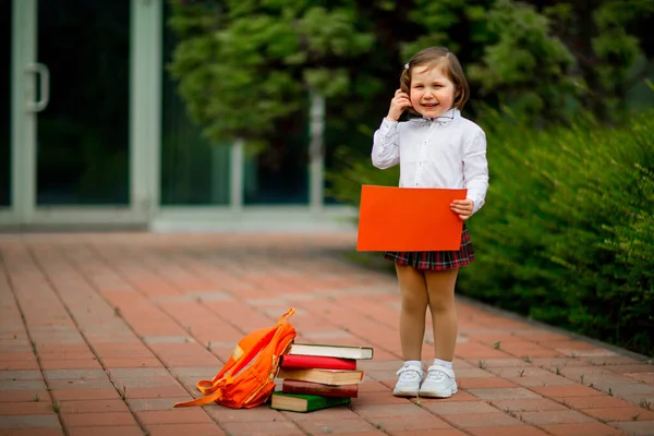 Een meisje in schooluniform staat vlak bij de school, met een stuk papier, een lay-out met ruimte voor tekst — Stockfoto