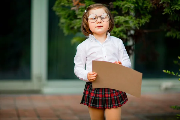 Una niña en uniforme escolar está cerca de la escuela, con un pedazo de papel, un diseño con espacio para el texto — Foto de Stock
