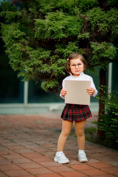 Een meisje in schooluniform staat vlak bij de school, met een stuk papier, een lay-out met ruimte voor tekst — Stockfoto