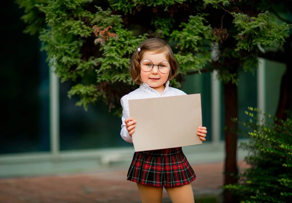 En flicka i skoluniform står nära skolan, med en bit papper, en layout med plats för text — Stockfoto