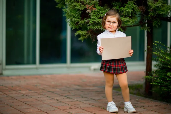 Uma menina de uniforme escolar fica perto da escola, com um pedaço de papel, um layout com espaço para texto — Fotografia de Stock