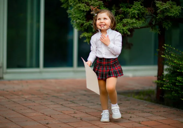 Uma menina de uniforme escolar fica perto da escola, com um pedaço de papel, um layout com espaço para texto — Fotografia de Stock