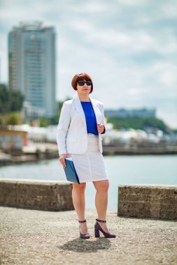 Güzel bir kadın, kahverengi saçlı, beyaz takım elbiseli ve mavi bluzlu, ellerinde belgelerle, deniz kenarında şehrin arka planında duruyor, bir iş portresi. .