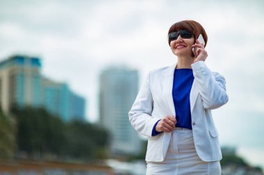Güzel bir kadın, kahverengi saçlı, beyaz takım elbiseli ve mavi bluzlu, ellerinde belgelerle, deniz kenarında şehrin arka planında duruyor, bir iş portresi. .