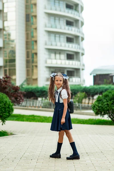 Красивая девушка в синей школьной форме, со школьным рюкзаком, рядом со школьным зданием. Возвращение в школу, 1 сентября — стоковое фото