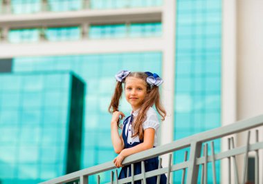 Mavi okul üniformalı, iki at kuyruklu bir kız öğrenci, okulun arka planında, cam duvarlarla duruyor..