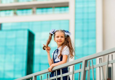 Mavi okul üniformalı, iki at kuyruklu bir kız öğrenci, okulun arka planında, cam duvarlarla duruyor..