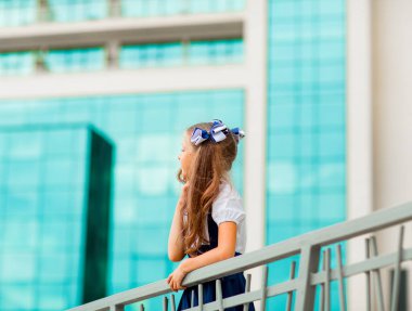 Mavi okul üniformalı, iki at kuyruklu bir kız öğrenci, okulun arka planında, cam duvarlarla duruyor..