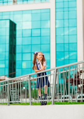 Mavi okul üniformalı, iki at kuyruklu bir kız öğrenci, okulun arka planında, cam duvarlarla duruyor..