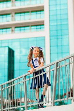 Mavi okul üniformalı, iki at kuyruklu bir kız öğrenci, okulun arka planında, cam duvarlarla duruyor..