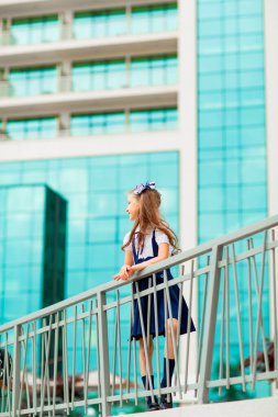 Mavi okul üniformalı, iki at kuyruklu bir kız öğrenci, okulun arka planında, cam duvarlarla duruyor..