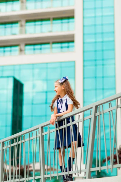 Mavi okul üniformalı, iki at kuyruklu bir kız öğrenci, okulun arka planında, cam duvarlarla duruyor..