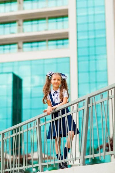 Mavi okul üniformalı, iki at kuyruklu bir kız öğrenci, okulun arka planında, cam duvarlarla duruyor..