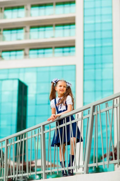 Mavi okul üniformalı, iki at kuyruklu bir kız öğrenci, okulun arka planında, cam duvarlarla duruyor..