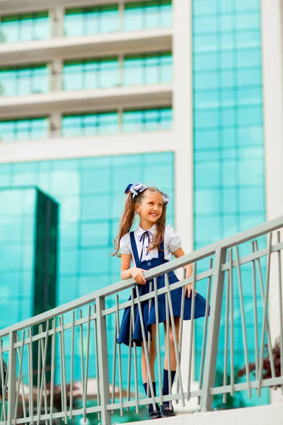 Mavi okul üniformalı, iki at kuyruklu bir kız öğrenci, okulun arka planında, cam duvarlarla duruyor..