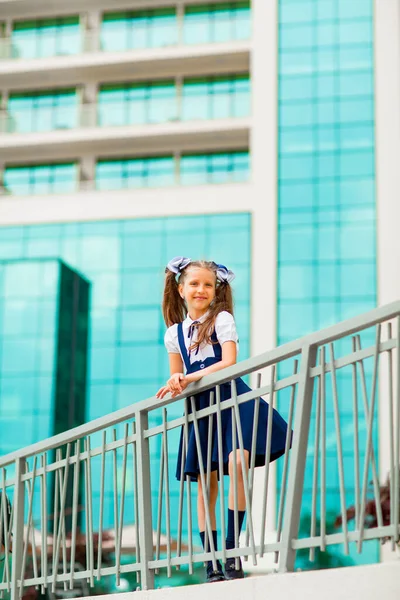 Mavi okul üniformalı, iki at kuyruklu bir kız öğrenci, okulun arka planında, cam duvarlarla duruyor..