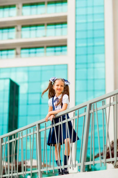 Mavi okul üniformalı, iki at kuyruklu bir kız öğrenci, okulun arka planında, cam duvarlarla duruyor..