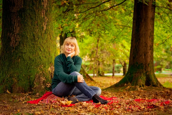 beautiful middle-aged woman,blonde,sitting under a large tree,on a red blanket of autumn leaves,on an autumn beautiful day