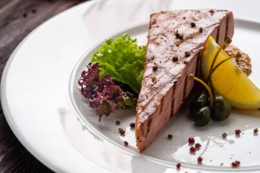 Fried orkinos biftek baharat ile yuvarlak beyaz plaka ve taze yeşil salata limon ve ahşap masa üzerinde Kapari.