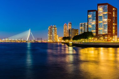 Illuminated Erasmus cable-stayed bridge over Maas river by buildings against blue sky at night, yellow lights reflected in the water. Rotterdam night cityscape in the Netherlands