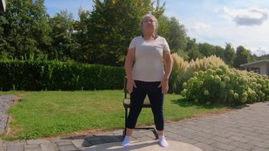 Aşırı kilolu yetişkin bir kadının bahçedeki sandalyeye çömelmesi, esnekliği artırmak için bacak ve dizlerini güçlendirmesi ön planda. Sağlıklı bir yaşam tarzı arayan yaşlılar için ev ödevi planı
